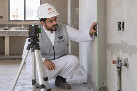 مهندس ميداني بالثوب وسترة الشركة الرمادية وخوذة بيضاء يتحقق من استقامة الجدار بميزان يدوي محاذٍ لخط ليزر أخضر في مطبخ قيد التجهيز.