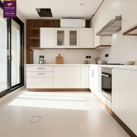 مطبخ أبيض ناصع بتوزيع زاوي L وخزائن علوية زجاجية بجوار نافذة شرفة مطلة.
