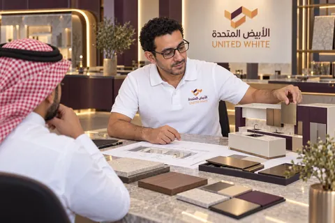 استشاري بقميص الشركة الأبيض يناقش عينة تشطيب معدني ومجسم مطبخ مع عميل، فوق طاولة تضم مخططات وعينات داخل صالة عرض عاجية وبنفسجية داكنة.