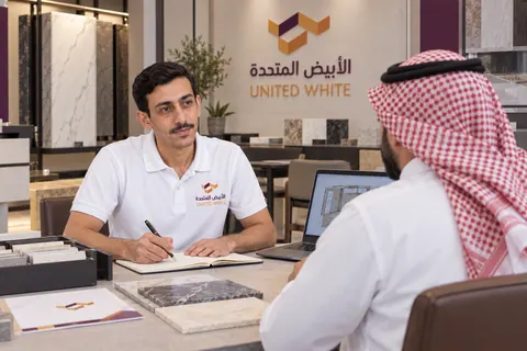 استشاري مبيعات بقميص الشركة الأبيض يستمع لعميل في صالة العرض ويدوّن متطلباته، بجوار عينات تشطيب وحاسوب محمول يعرض مخطط المطبخ.