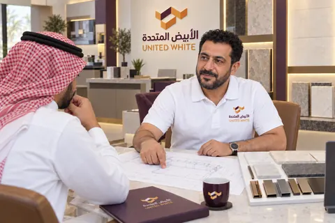 استشاري بقميص الشركة الأبيض يراجع مخططاً هندسياً مطبوعاً للمطبخ مع عميل، وسط عينات معدنية وحجرية وملف توثيق الشركة على طاولة الاستشارة.